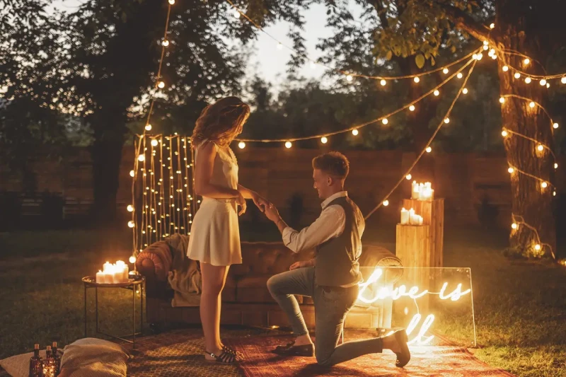 Un homme agenouillé fait sa demande en mariage dans un jardin décoré de guirlandes lumineuses, bougies et un panneau « Love is all », créant une ambiance magique.